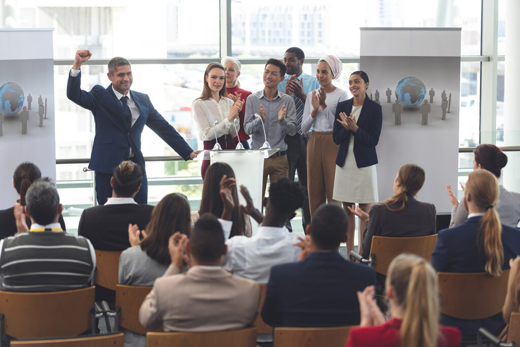 Confident business leader speaking at podium with diverse team applauding during corporate leadership event